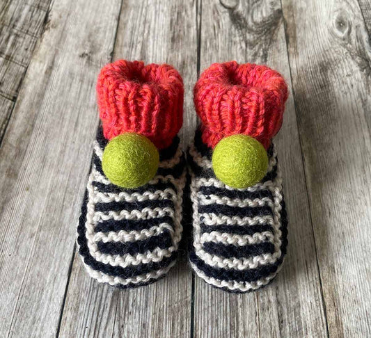 Pair of hand knitted knitted striped baby booties with red tops and green pom-poms on a wooden surface made by lilly and bright