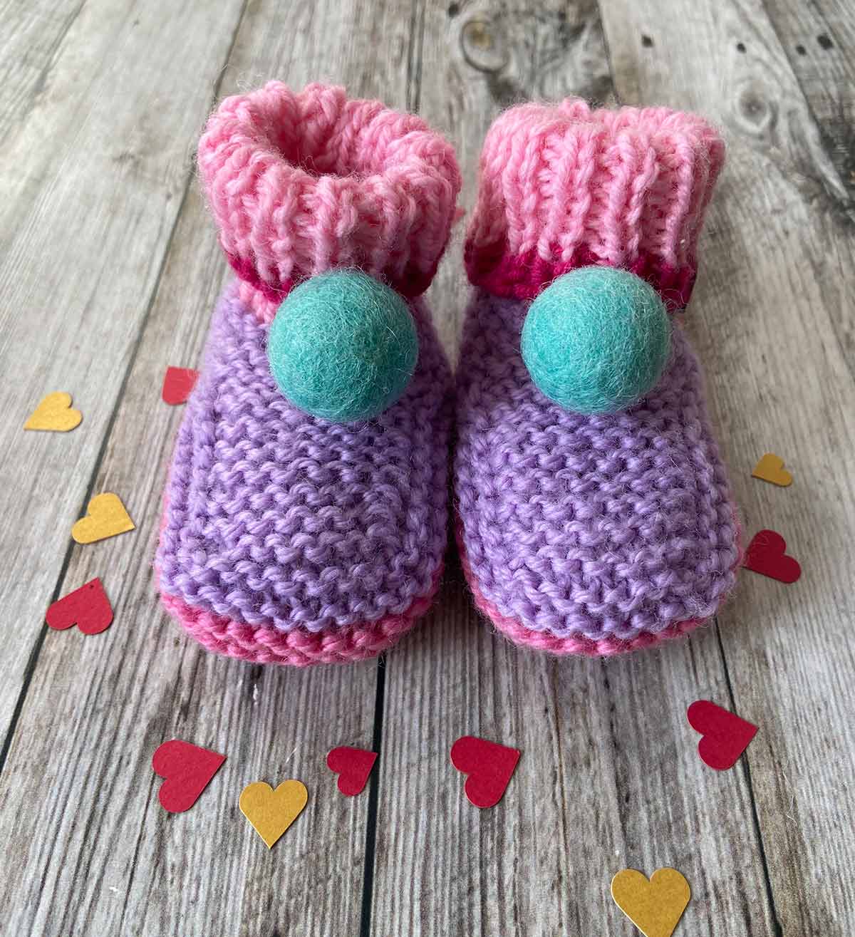 Pair of pink and purple hand knitted baby booties with teal pom-poms on a wooden surface with heart-shaped confetti. Made by lilly and bright