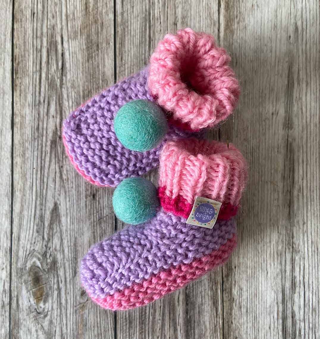 Pair of hand knitted lilac baby booties with aqua blue pom-poms on a wooden surface - maade by lilly and bright