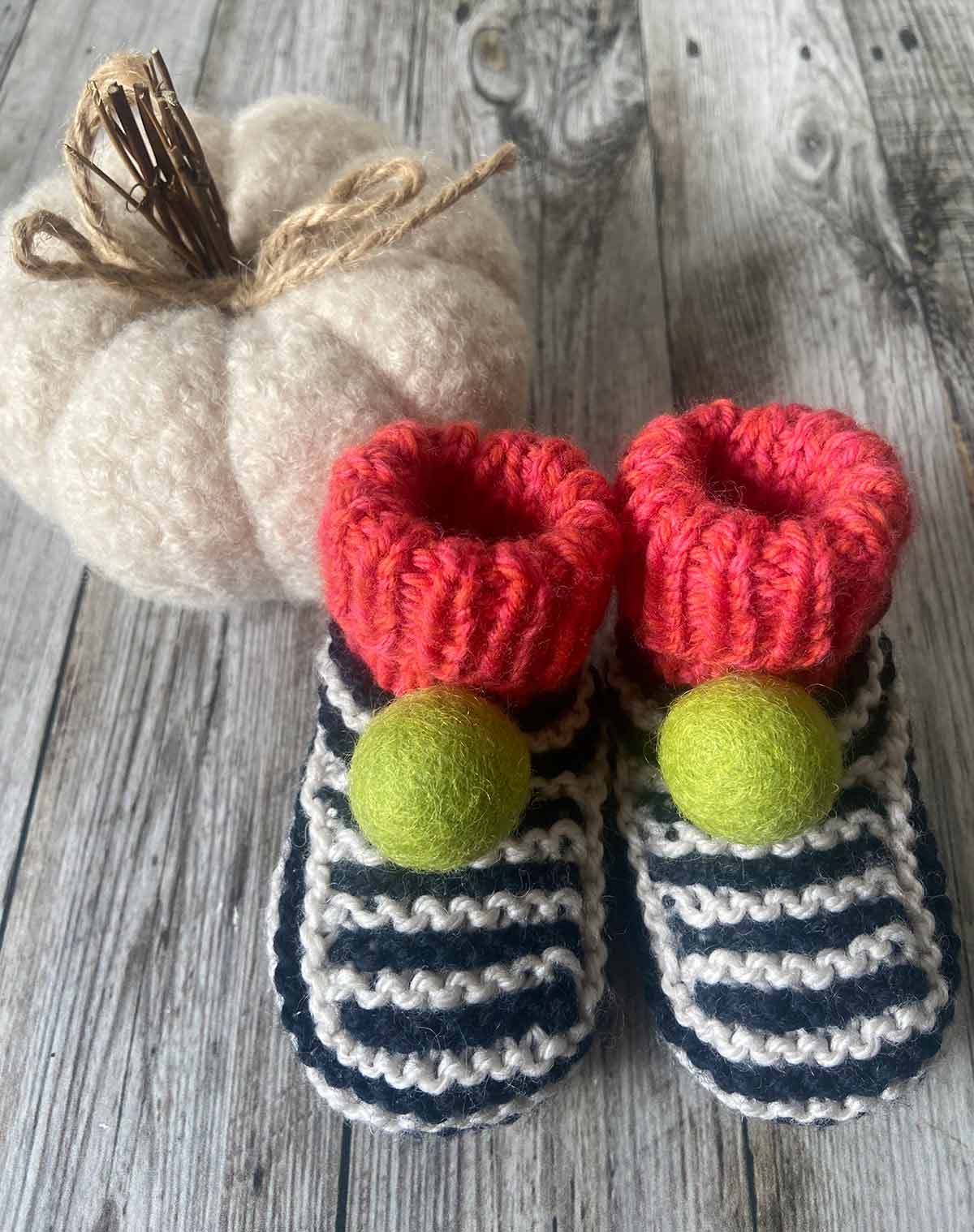 Pair of hand knitted striped baby booties with red tops and green pom-poms on a wooden surface with a pumpkin - by lilly and bright