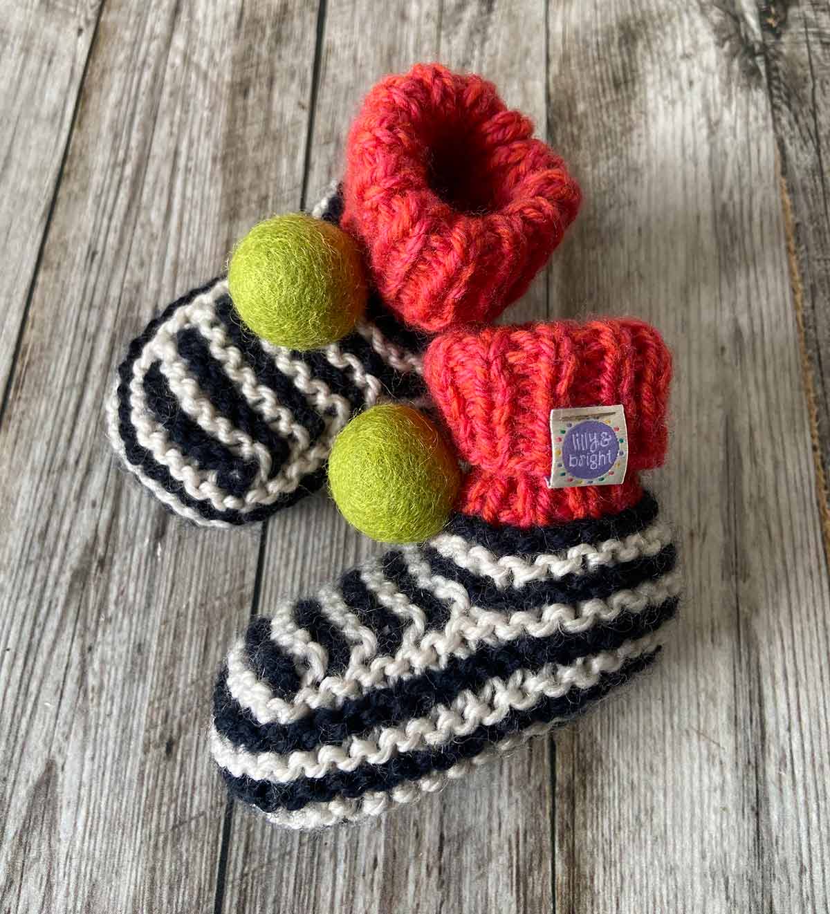 Pair of hand knitted striped baby booties with red tops and green pom-poms on a wooden surface by lilly and bright