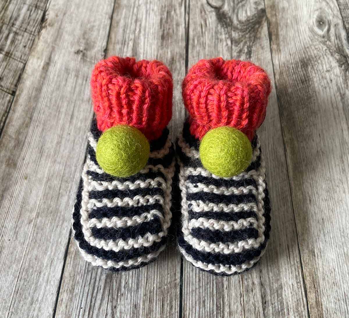 Pair of hand knitted knitted striped baby booties with red tops and green pom-poms on a wooden surface made by lilly and bright
