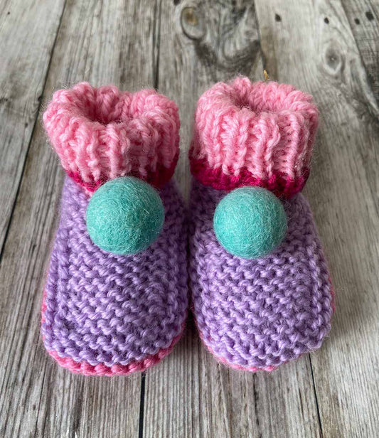 Pair of hand knitted knitted baby booties with pink tops and purple bodies with a blue pom pom made by lilly and bright